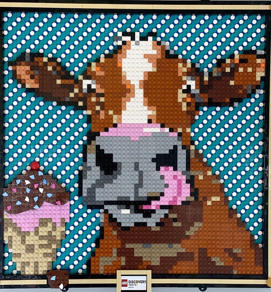 Mural made of LEGO pieces a brown and white spotted cow face with a grey nose and a pink tongue sticking out Lower left corner has a cone of pink icecream covered in hot fudge and a cherry Blue and white background All comprised of LEGO pieces