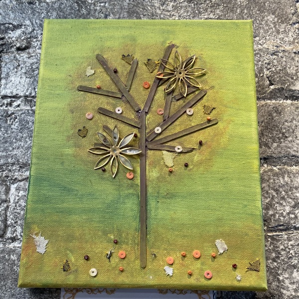 Painted green canvas with a brown stick tree centered Darker green halo around tree suggesting canopy with two gold flowers glued into the canopy along with small orange and silver small buttons Below tree is suggestion of grass the same color as the ca