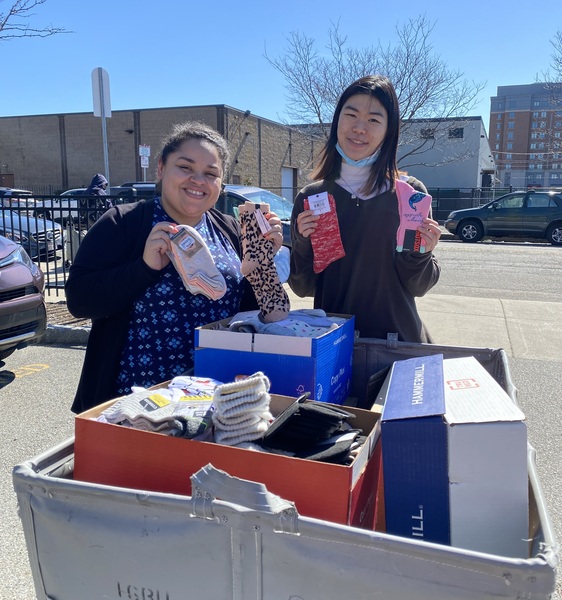 Two women holding new pairs of socks standing behind a white hamper filled with boxes of new pairs of socks. Blue skies behind them. 