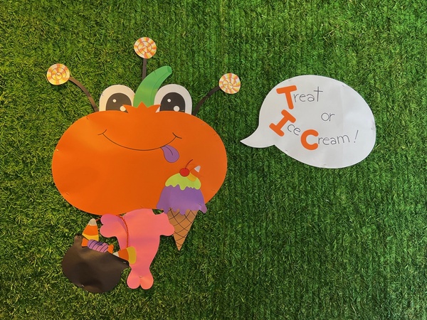 Art made of cardboard cut outs. A round orange face that039s actually a pumpkin with two large white, round eyes on top of the pumpkin with three round antena. The creature has a small, pink body and is holding an ice cream cone of purple and green ice