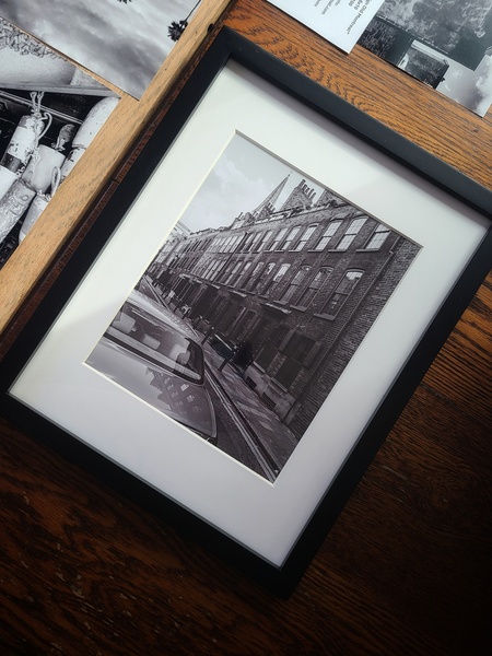 Photograph of a framed photograph It is a black and white photo of a street of brownstone buildings in a black frame on a wooden table shot from above You can just make out that there are unframed photos on the floor below that table