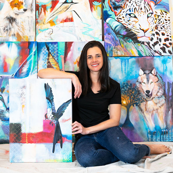 Artist a woman in a black shirt jeans and barefoot with long black hair leaning on a painting of a bird in front of a wall of colorful paintings of a wolf a leopard a fox and more that are slightly out of view