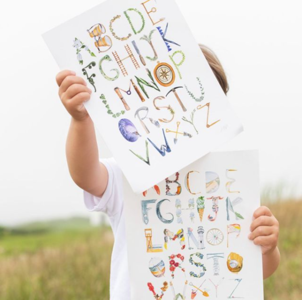 Young child holding two pieces of artwork framed in white frames Face and body is blocked by frames The art is the alphabet with each letter a piece of decorative art Standing in a green field 