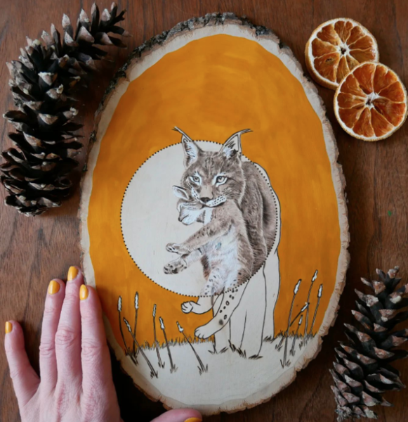 Oval of wood with bobcat carrying baby burned onto piece Orange painted background with left hand in lower left corner holding piece in place nbsp