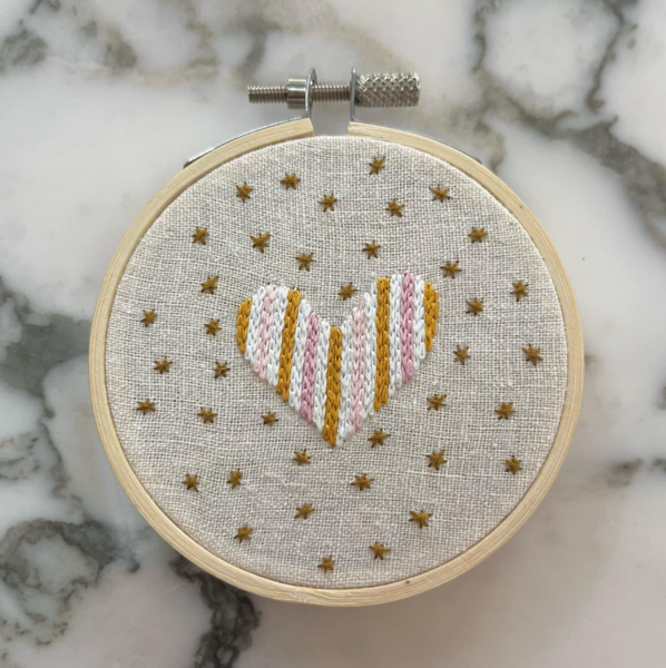 Small embroidery hoop with off white fabric, light brown stars and pink, white, and light brown heart embroidered in middle. On marble surface. 