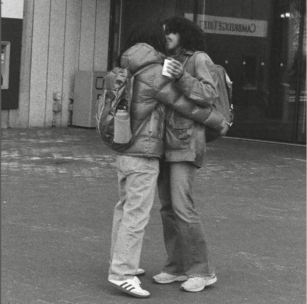 Black and white photograph Two people with dark shoulder length hair embracing on a brick sidewalk in front of a concrete building Both are wearing backpacks and jeans and white sneakers one is holding coffee in their left hand You can just see a smi