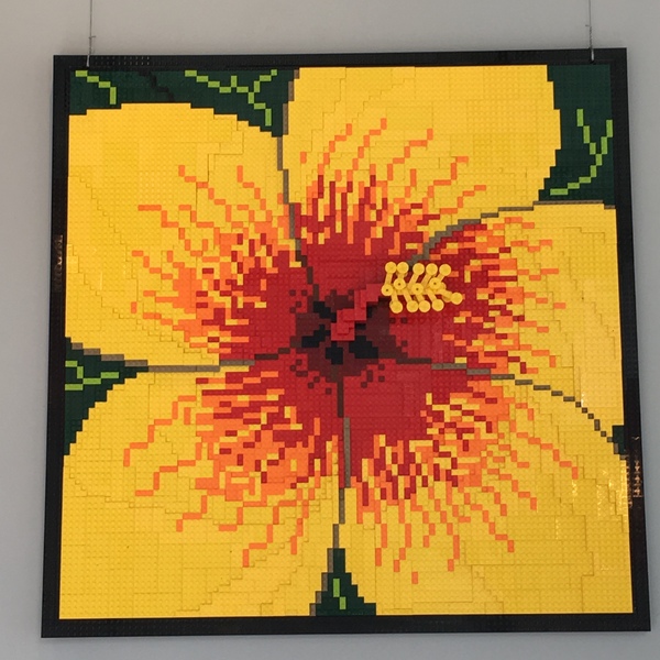 Mural of LEGO pieces hanging on wall Looks like a yellow Hibiscus with red center with black border Completely made of LEGO pieces 