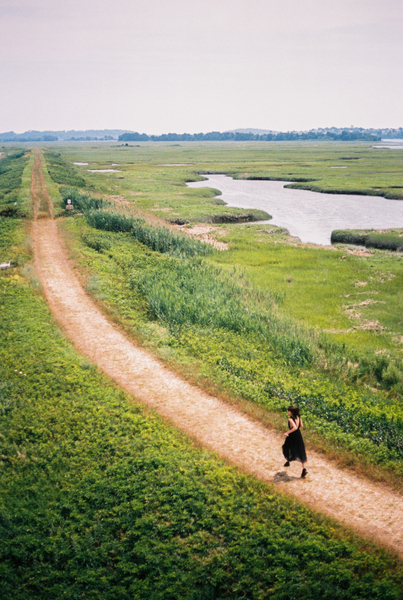 Photograph of a light brown dirt path cutting through a field of green grass There is a small pond in the upper right quadrant of the picture A long figure wearing black strides away from us in the lower right quadrant of the picture The sky is a blue 