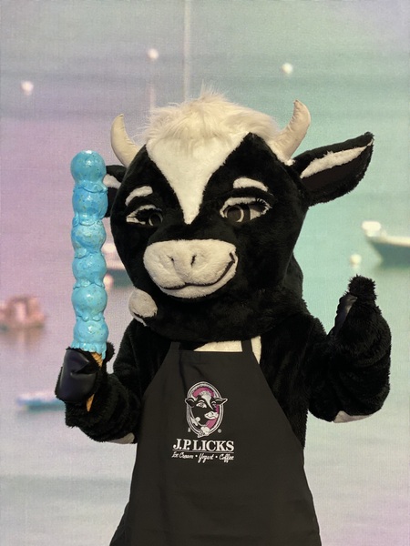 JP Licks cow mascot Black and white spotted cow with white horns and a tuft of white hair on the top of her head She is wearing a black apron with the JP Licks logo She is holding a fake stack of blue ice cream on a fake cone in front of a pastel p