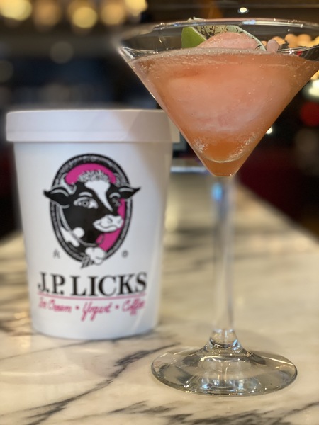 Covered white JP Licks branded quart container sitting slightly behind and to left of a martini glass filled with blended pink sorbet and vodka and half of charred lime a drink called For Petes Sake All sitting on a white and grey m