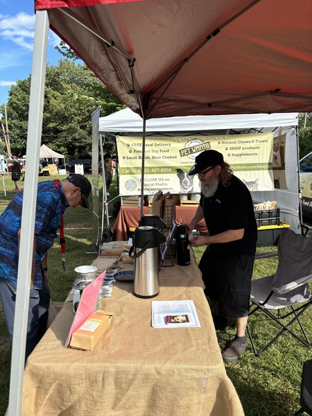 Man with long grey beard wearing all black including black hat socks and shoes pouring cup of coffee out of a carafe to an older gentleman wearing all blue over a table covered in burlap underneath a red tent open on all four sides 