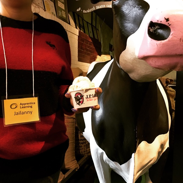 Torso of person in red and black striped sweater wearing name tag from Apprentice Learning holding JP Licks cup of ice cream next to ceramic cow