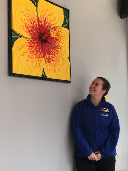 Artist standing under and looking up at a mural made completely of LEGOS. Mural looks like a hibiscus with yellow petals and red center surrounded by black border.