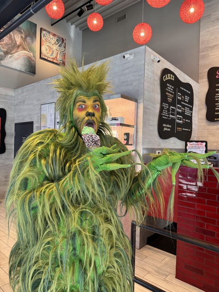 Man in Grinch costume green costume shrugging while holding the Grinchs Candy Cane Ice cream in peppermint dipped cone at Assembly Rows JP Licks store Facial expression is eyes widened and licking ice cream