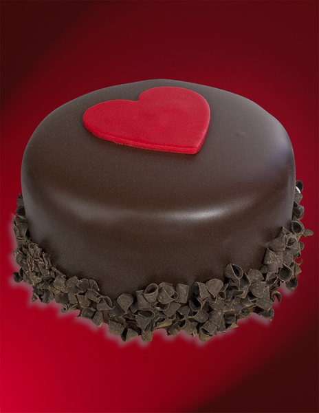 Chocolate covered ice cream cake with chocolate shavings along bottom and red fondant heart on top on red background 