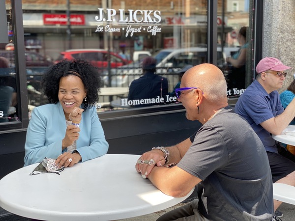 Mayor Janey in blue jacket sitting at white table with chocolate ice cream in a cone smiling and looking off camera with owner Vincent Petryk in black shirt sitting across from her as seen from the left side