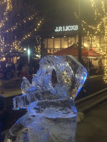 Ice sculpture of hooded figure in front of JP Licks with lights in trees in the middle ground 